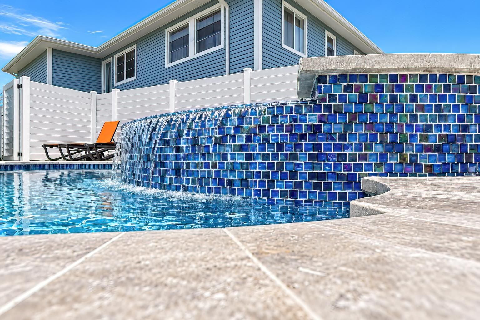 A swimming pool with a mosaic tile wall, a lounge chair, and a house in the background.