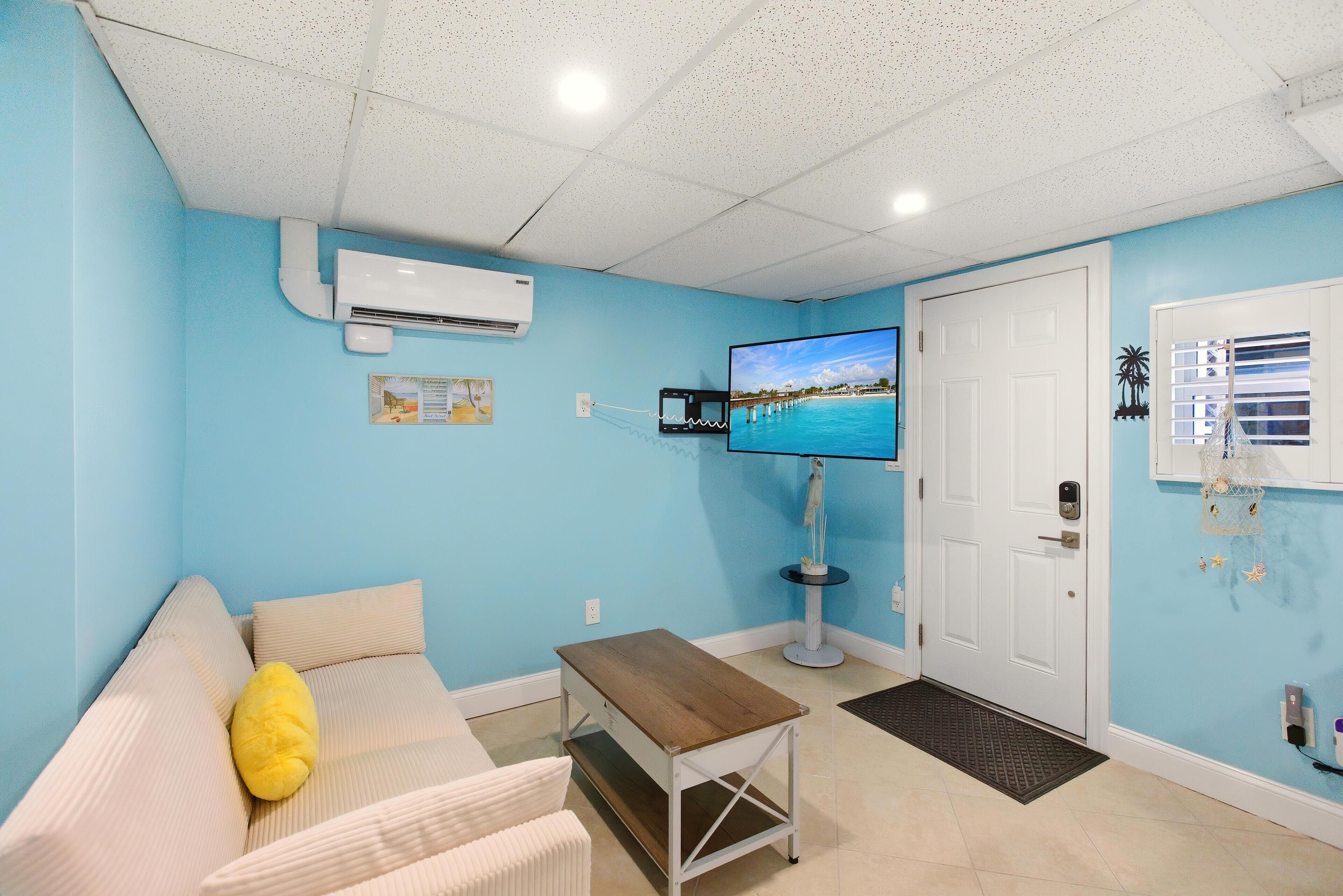 A room with a light blue wall, a white door, a television, a small table, and a sofa with a yellow pillow.