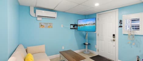 A room with a light blue wall, a white door, a television, a small table, and a sofa with a yellow pillow.