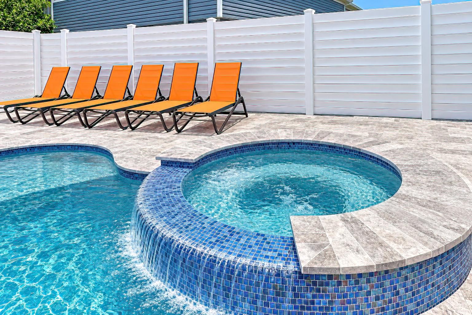 Piscina com borda revestida, cercada por um muro branco e cadeiras de descanso laranja.