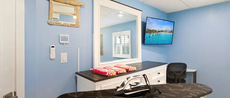A room with a large mirror, a television, an ironing board, and a desk with a chair.
