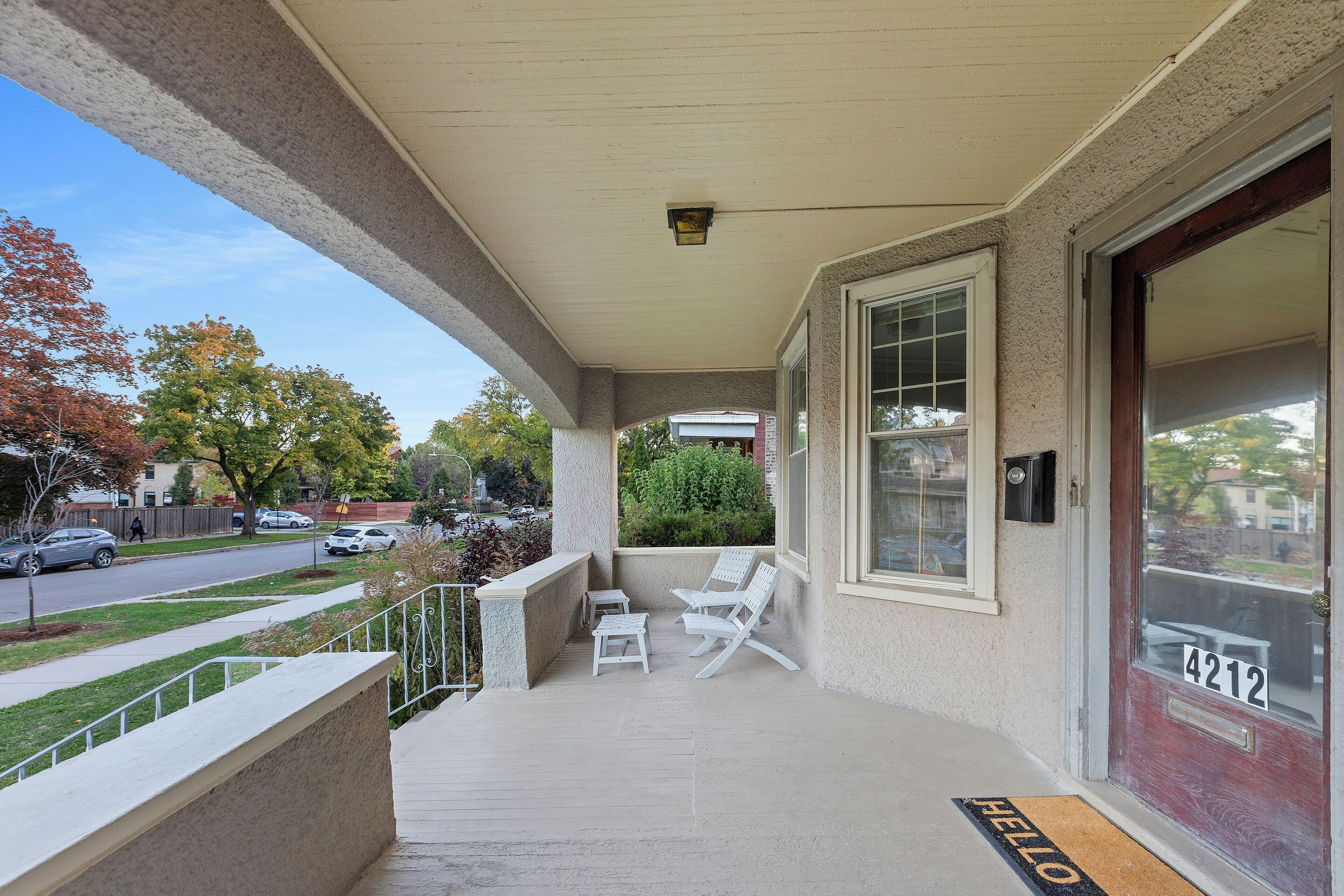 A covered porch with two white chairs and a small table, a red door with the number 4212, and a 'HELLO' mat at the entrance.