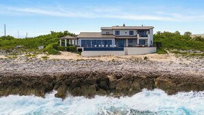 Une maison moderne à deux étages, dotée d’un balcon, située sur une plage rocheuse où les vagues déferlent à proximité.