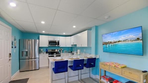 A modern kitchen with white cabinets, a stainless steel refrigerator, and a microwave. There is a dining area with blue chairs and a wooden console table. - Coral Suite (Fort Myers Beach)
