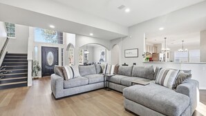 A spacious living room with a grey sectional sofa, a staircase, and a kitchen area. - Luxury Backyard Pool & Patio Oasis (4-BD/2.5-BA) (Lewisville)