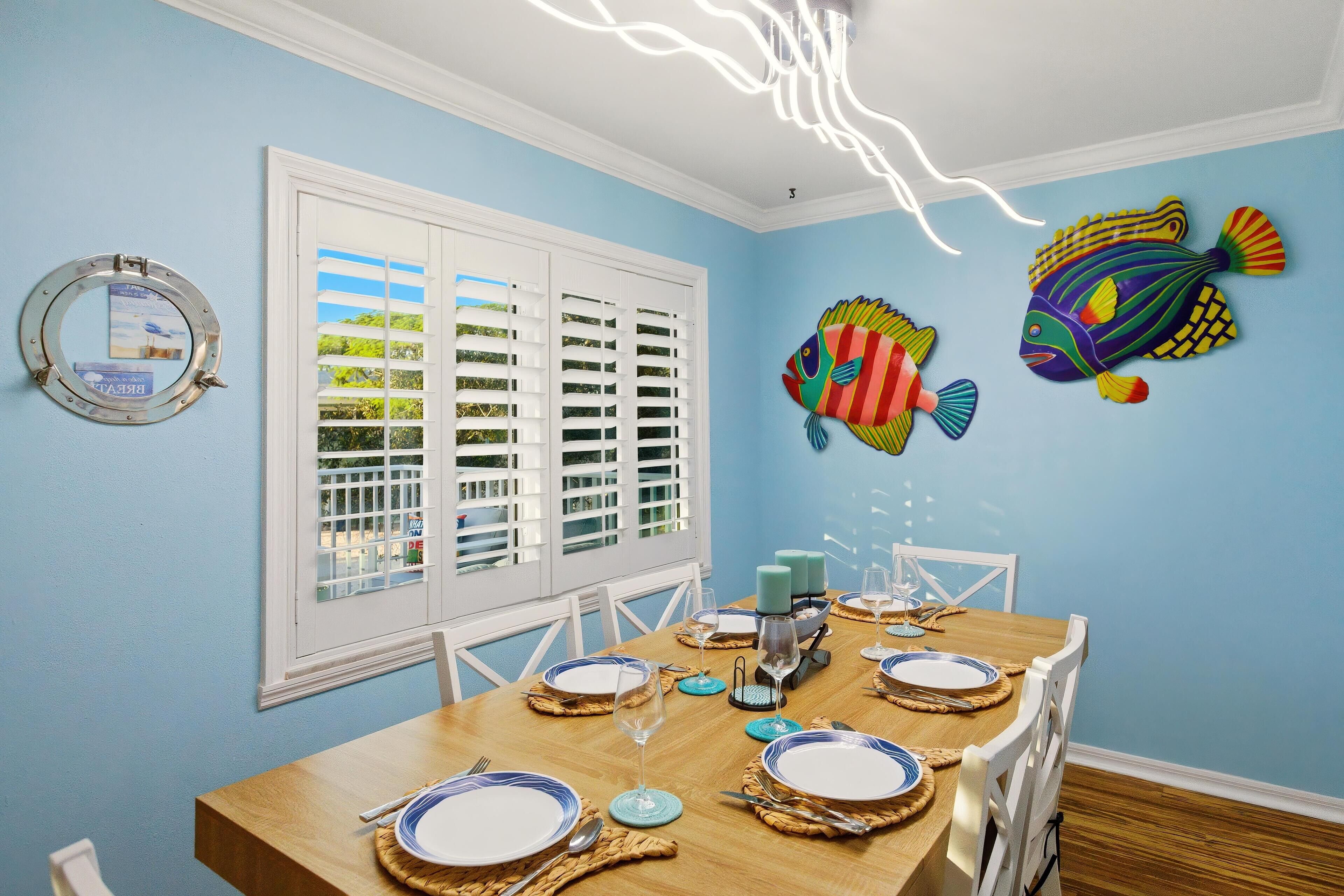 A dining area with a wooden table set for four, white chairs, and a view through the window with white shutters.
