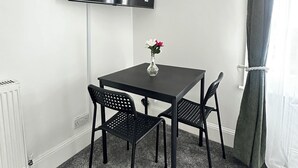 Ein kleiner Raum mit einem schwarzen Tisch und zwei Stühlen, einem flachbildfernseher an der Wand, einem Heizkörper und einem fenster mit Vorhängen.