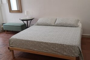 Ein ordentlich bezogenes Bett mit gemustertem Bettbezug und Kissen, ein kleiner runder Tisch mit einer Lampe sowie ein grüner Hocker in einem schlichten Raum mit Fliesenboden.