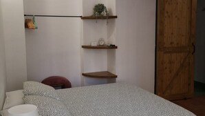 A bedroom with a bed, a red chair, and wooden shelves with a clock and a plant. - Salvia is an independent loft located on the ground floor, featuring a small private courtyard furnished with chairs and a table. The space consists of an open-plan area with a sofa bed and equipped kitchen, as well as a spacious bathroom with a shower.  (Santo Stefano di Camastra)