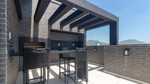 Une terrasse extérieure avec un mur en briques, un barbecue, deux tabourets de bar et une structure en forme de pergola.