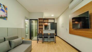A modern living room with a grey sofa, a dining area with three chairs, a kitchen with a TV, and a hallway leading to a bathroom.