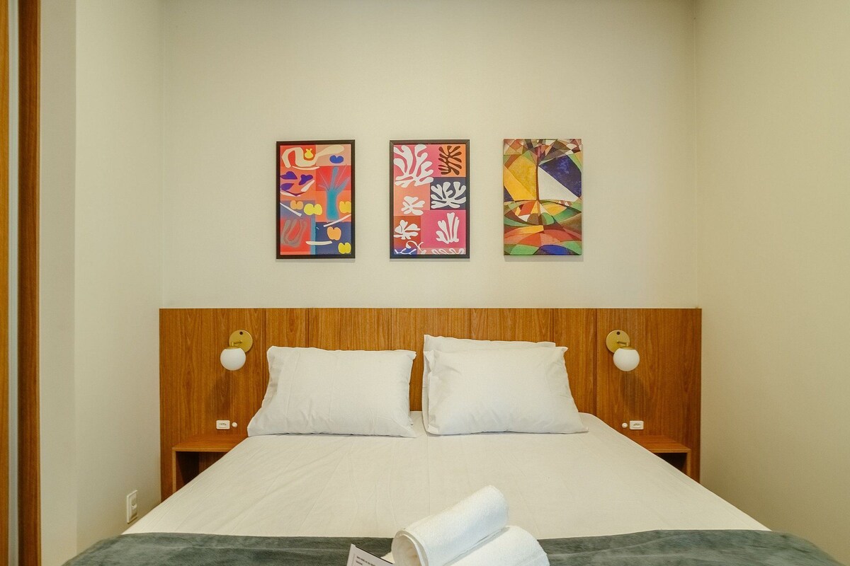 A hotel room with a wooden headboard, two white pillows, a bed with a white bedspread, and three abstract paintings on the wall.