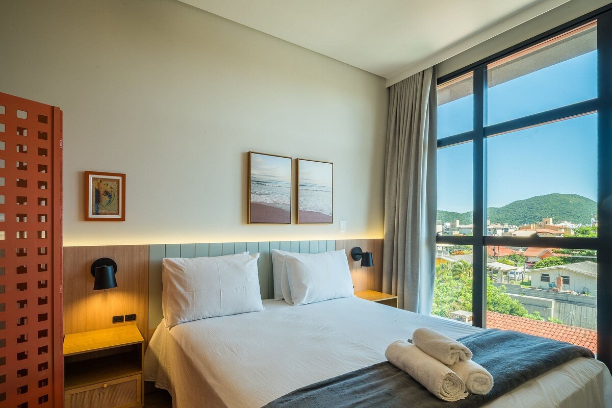 Hotelkamer met een bed, twee wandlampen, een raam met uitzicht op een stadsgezicht en een rode scheidingswand met een ruitpatroon.