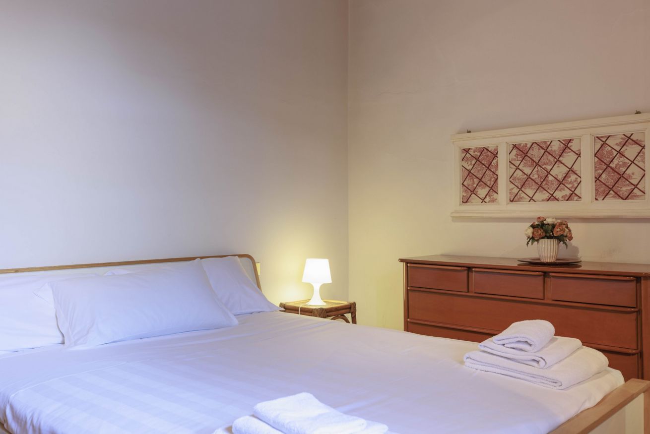 Una cama bien hecha con ropa blanca y toallas, una lámpara de noche, un aparador de madera y un panel decorativo sobre el aparador.