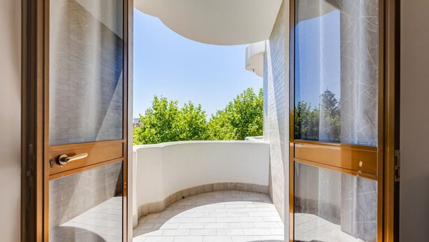 A balcony with a curved white wall, a door with a handle, and a clear view of green trees in the background. - BDC - Aurelia Seaview! (Santa Marinella)