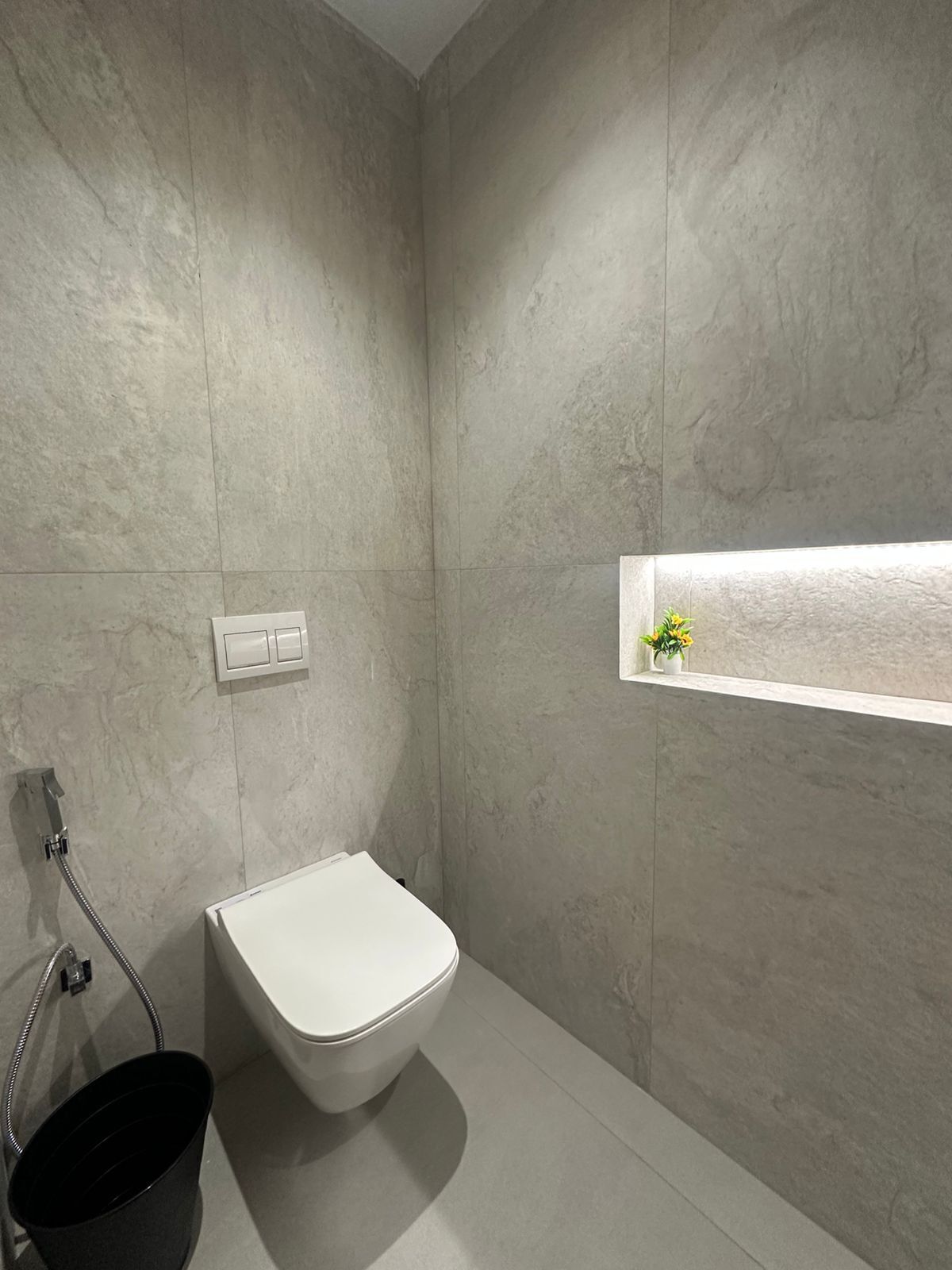 A modern bathroom with a white toilet, a black bucket, and a small shelf with a plant.