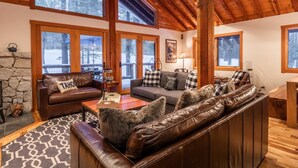 A cozy living room with a stone fireplace, leather sofas, and large windows offering a view of the snowy outdoors. - West Shore Home with Private Hot Tub (Tahoe City)