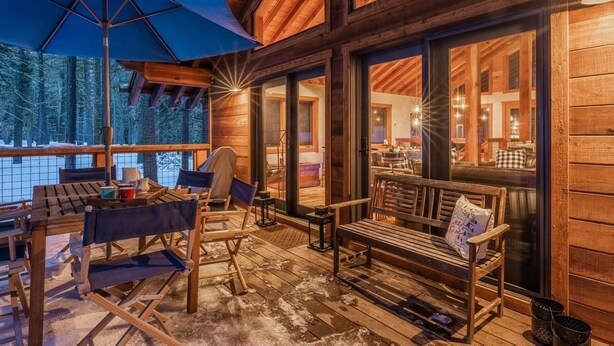 A wooden deck with outdoor seating, large windows, and a view of the surrounding forest. - West Shore Home with Private Hot Tub (Tahoe City)