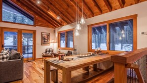 A cozy wooden interior with a dining table, chairs, and a sofa. Large windows offer views of a snowy landscape. - West Shore Home with Private Hot Tub (Tahoe City)
