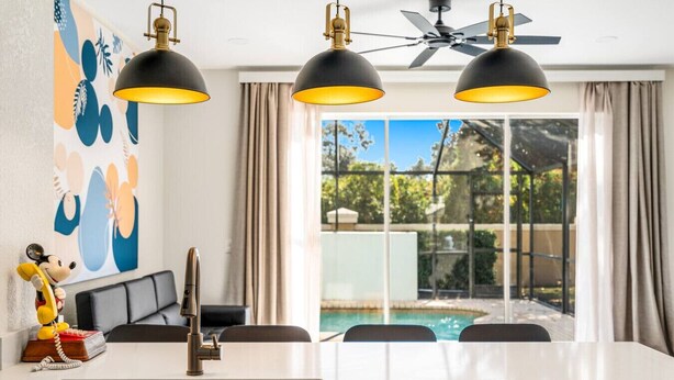 Un'area pranzo moderna con tavolo bianco, lampade pendenti nere, ventilatore a soffitto e vista su una piscina esterna.