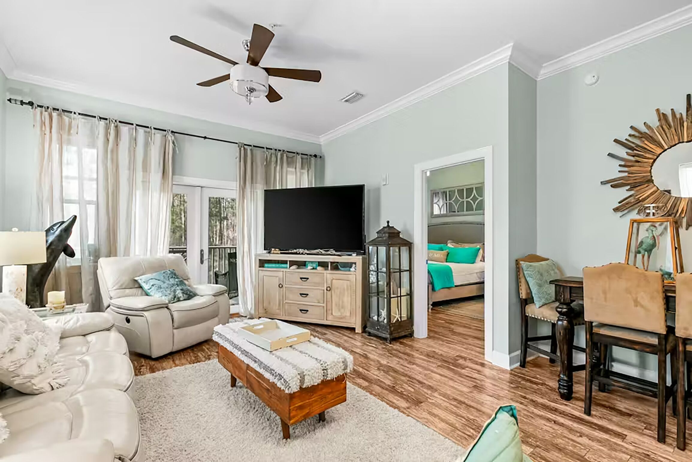 A spacious living room with a ceiling fan, a flat-screen TV, a dining area with wooden chairs, and a cozy seating area with a white sofa and ottoman.