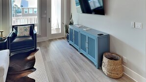 A modern living room with a flat-screen TV mounted on the wall, a blue cabinet, a leather sofa, and a wicker basket. - Serenity at Port A (Port Aransas)