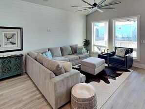 A modern living room with a sectional sofa, a small ottoman, a decorative cabinet, and a ceiling fan. - Serenity at Port A (Port Aransas)