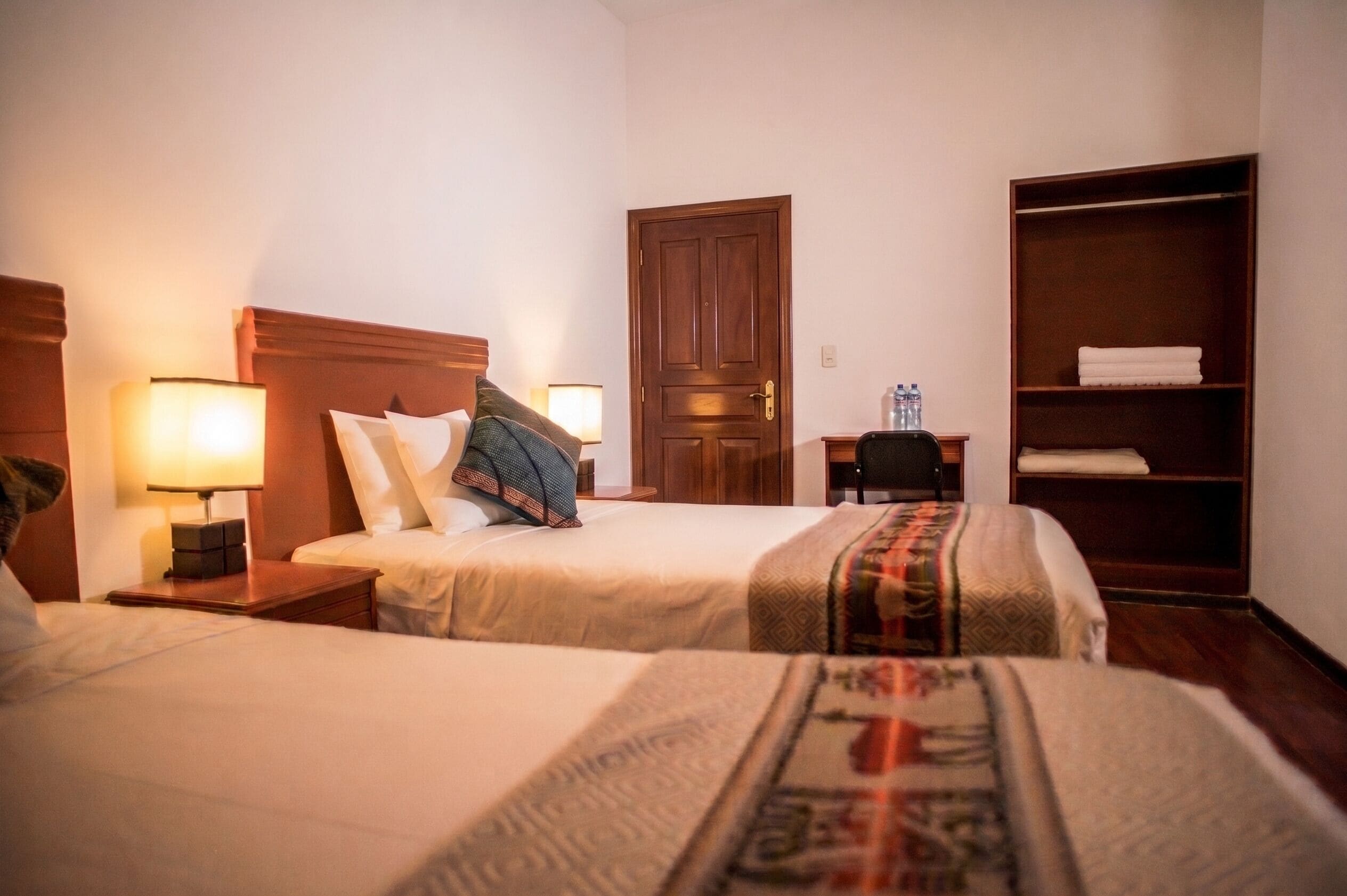 A hotel room with two beds, a wardrobe, a desk with a chair, and a door.