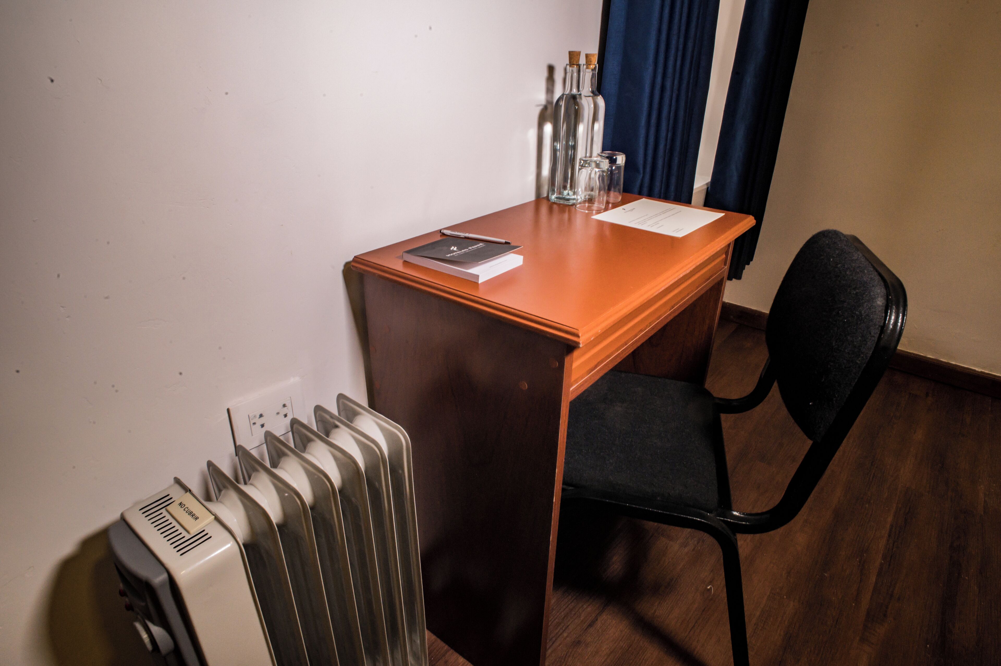A small wooden desk with a chair, a radiator, and a window with curtains.