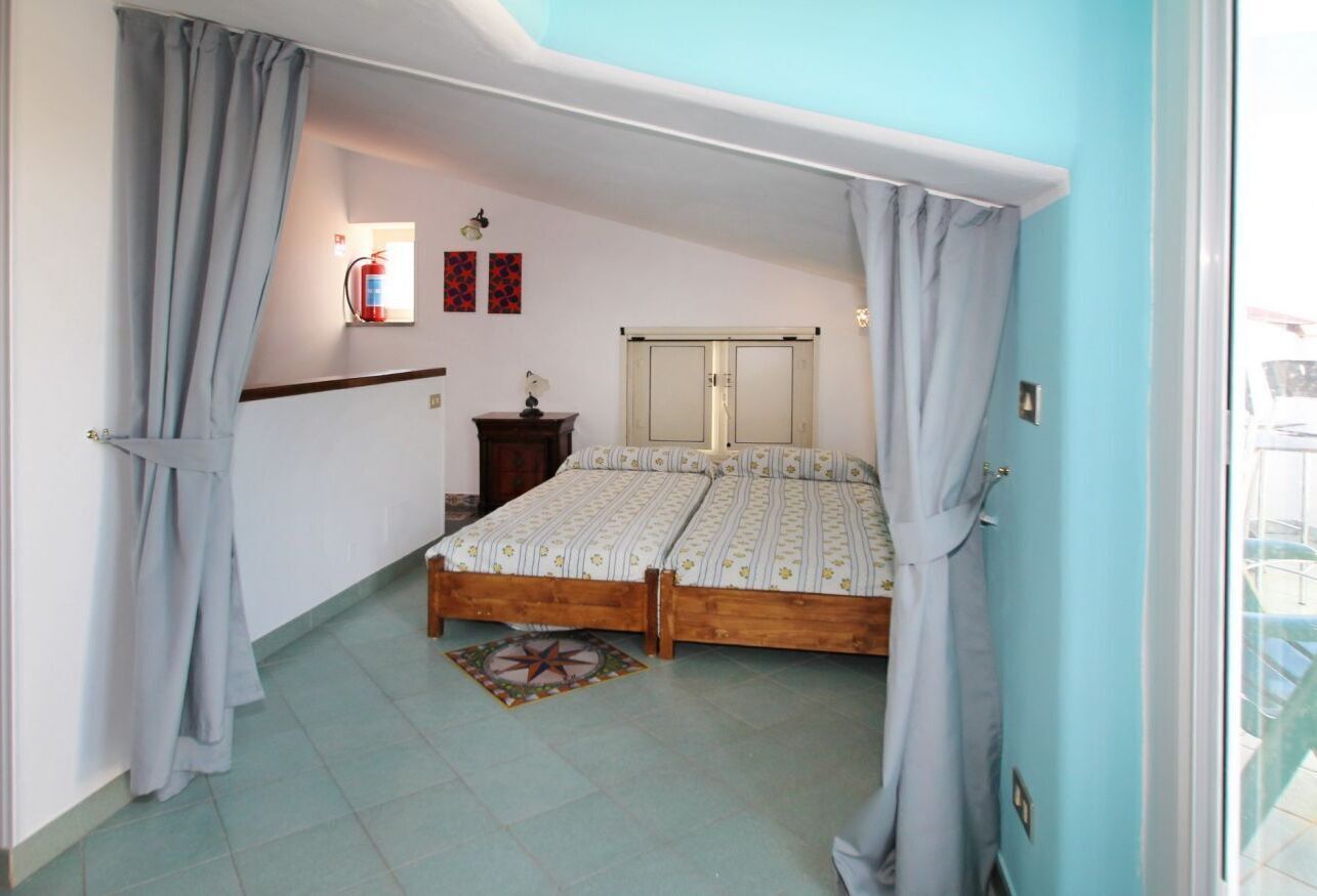 A bedroom with two beds, a small bedside table, a nightstand, a window with curtains, and a wall-mounted fire extinguisher.