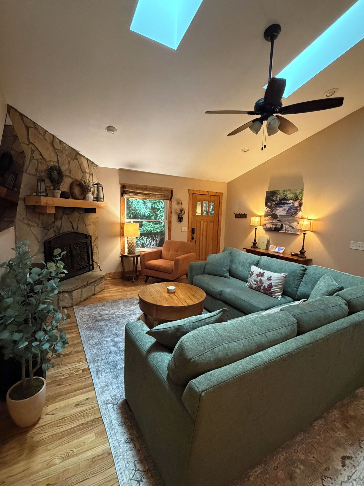 A cozy living room with a stone fireplace, a ceiling fan, and a large window.