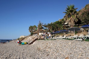 Ein Kiesstrand mit Liegestühlen und Sonnenschirmen, Palmen und einem klaren Himmel.