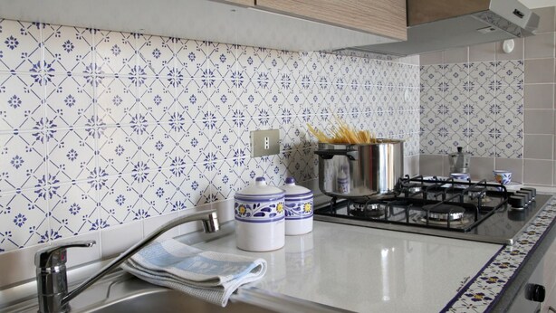 Una cucina con zoccolo piastrellato, lavello in acciaio inox e piano cottura a gas.