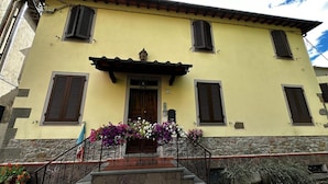 A two-story building with a yellow facade, stone base, and a wooden door. There are four windows with shutters, a small porch with steps, and a stone wall. - Turquoise fairy family suite (Collodi)