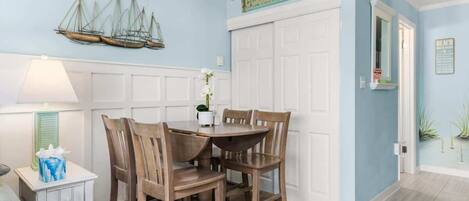 Un coin repas avec une table et des chaises, un bateau décoratif accroché au mur et une enseigne sur laquelle on peut lire « MAISON DE PLAGE ».