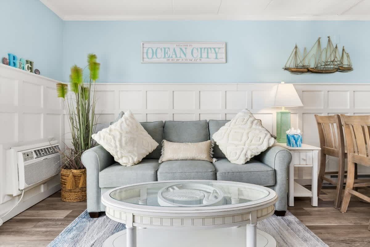 Ein Wohnzimmer mit grauem Sofa, einem Glastisch, einem Korbwäsches und einem Wandschild mit der Aufschrift 'Welcome to Ocean City'.
