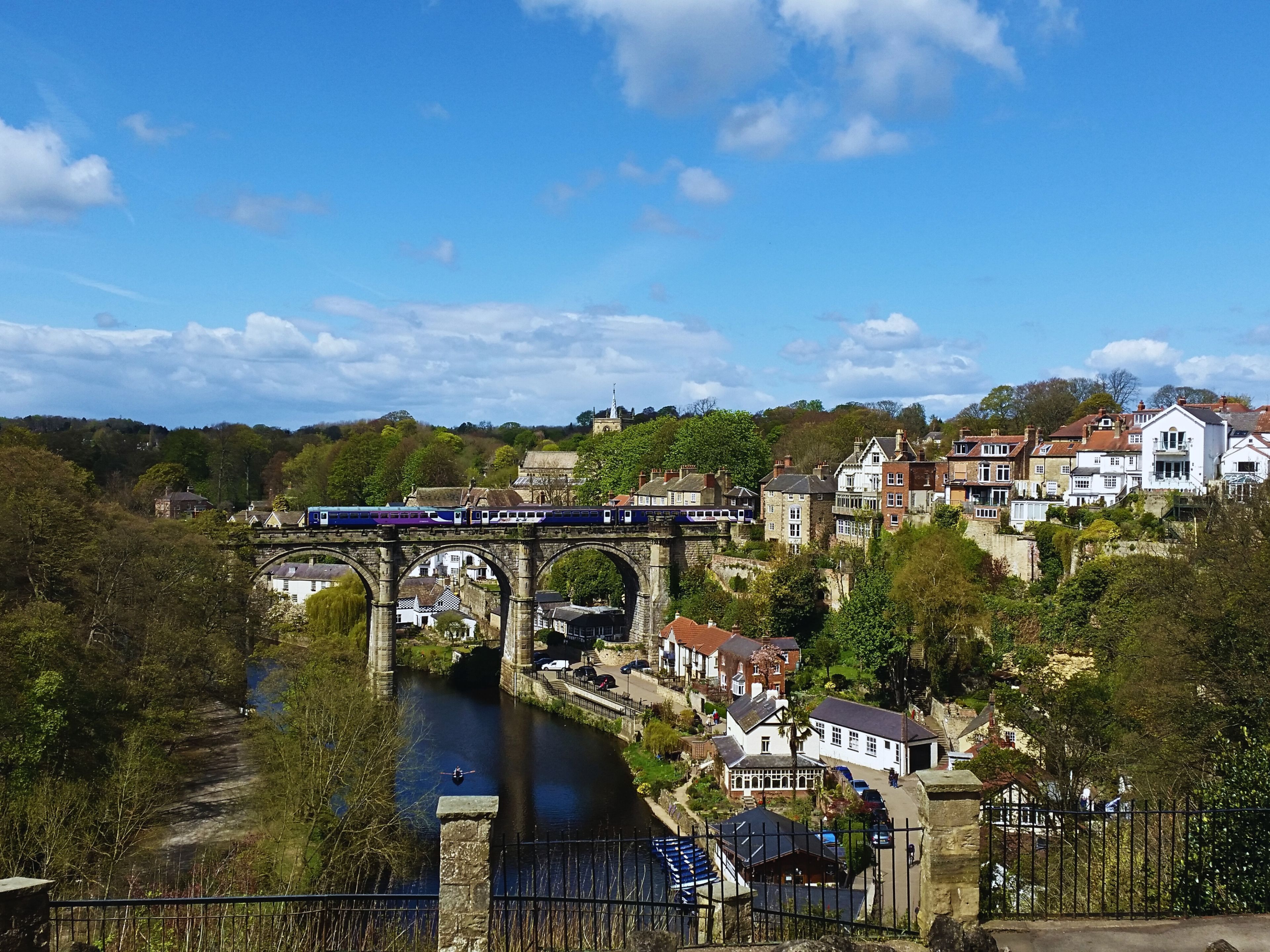 A train crossing a stone bridge over a river, with buildings and greenery in the background.