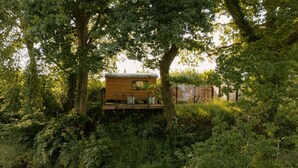 A wooden cabin with a deck, surrounded by dense greenery and a stream. - Dog friendly glamping accommodation with hot tub in Ripon, Yorkshire (West Tanfield - Ripon)