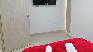 Une pièce avec un lit rouge, une porte en bois, un mur blanc et un téléviseur fixé au mur.