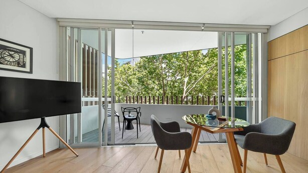Een moderne woonkamer met een flatscreen televisie op een driepoot, een glazen tafel met stoelen en een schuifpui naar een terras met uitzicht op bomen.