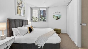 Een moderne slaapkamer met een groot bed, een nachtkastje met een lamp, een klein krukje, een raam met uitzicht op groen, en een ronde spiegel aan de muur.