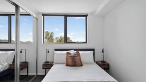 A modern bedroom with a large bed, two bedside tables, a mirror, and a view of the outdoors through large windows. - Northern Breeze Terrace Haven near Curl Curl Beach (Brookvale)
