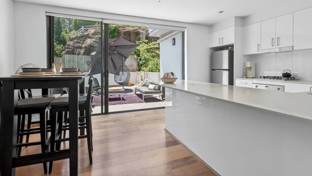 A modern kitchen with white cabinets, a large island, and a sliding glass door leading to a patio with outdoor furniture. - Northern Breeze Terrace Haven near Curl Curl Beach (Brookvale)