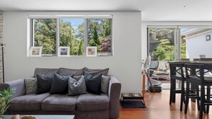 A modern living room with a grey sofa, a dining table with chairs, and a large window with framed pictures. - Northern Breeze Terrace Haven near Curl Curl Beach (Brookvale)