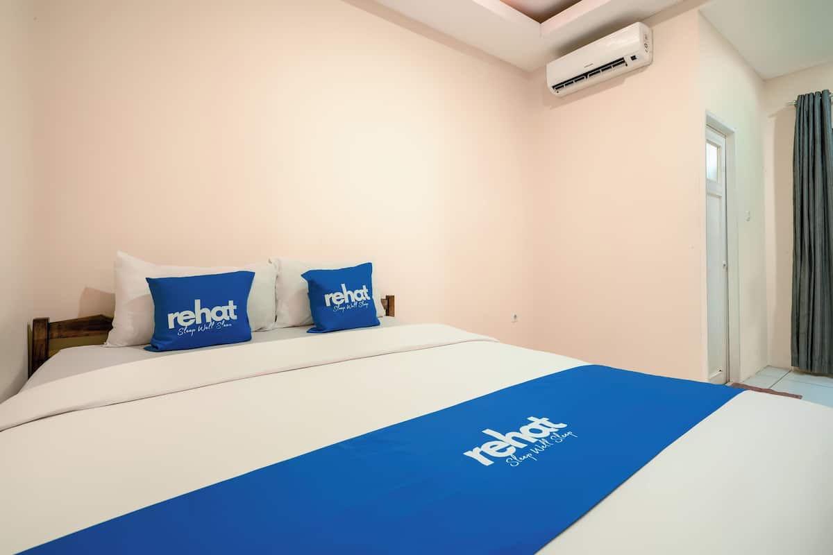 Une chambre d’hôtel avec un lit, deux oreillers portant la marque « rehat », une couvre-lit bleue et un climatiseur fixé au mur.