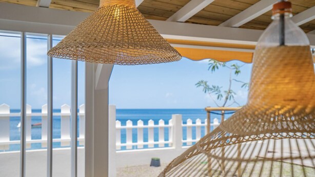 Ein Zimmer mit großen Fenstern mit Blick auf einen Strand, ausgestattet mit Korbstühlen und einem weißen Zaun.