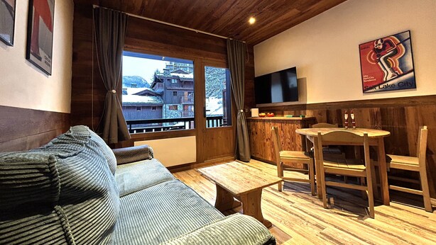 A cozy room with a wooden ceiling, a sofa, a dining table with chairs, a TV, and a window with a view of buildings. - Chalet de Solaise B03 (Val-d'Isère)