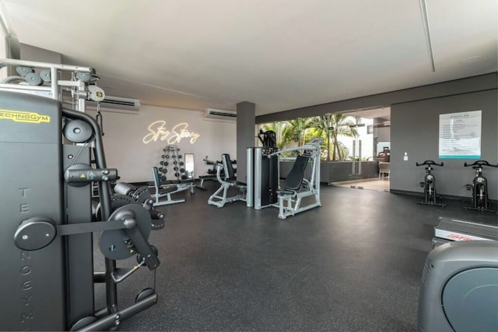 A gym with various exercise machines, a neon sign, and a view of palm trees outside.
