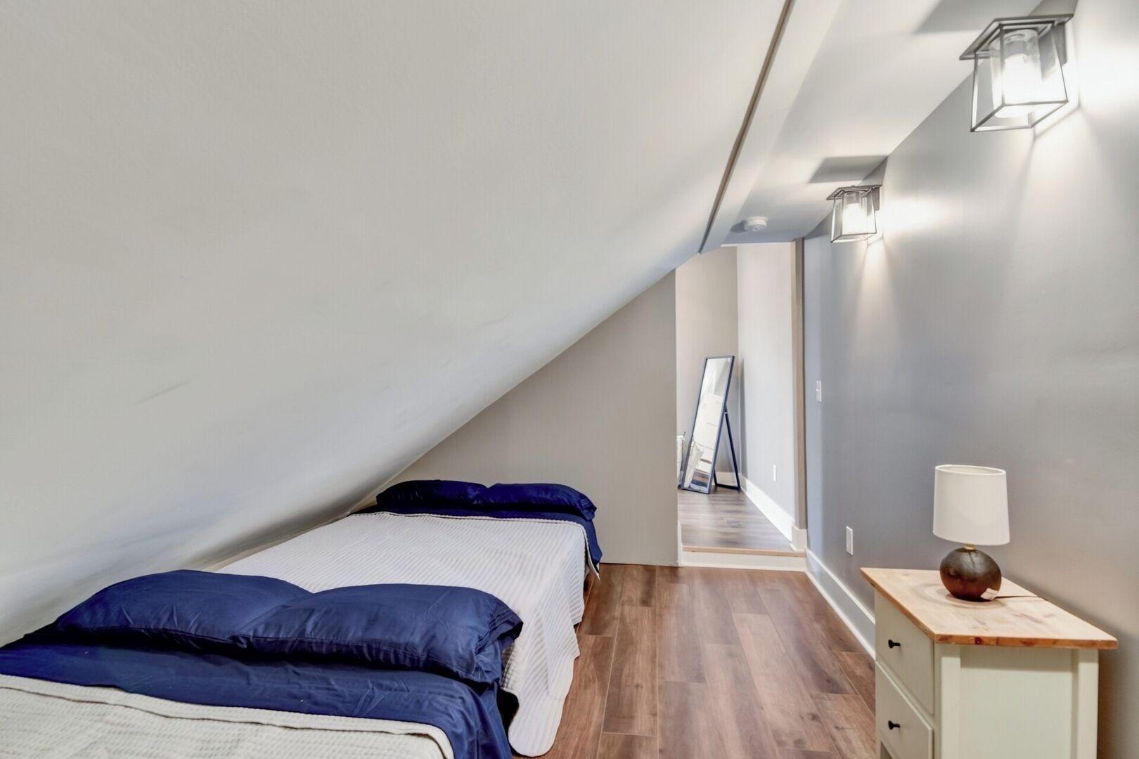 A bedroom with a sloped ceiling, a bed with blue bedding, a bedside table with a lamp, and a mirror leaning against the wall.