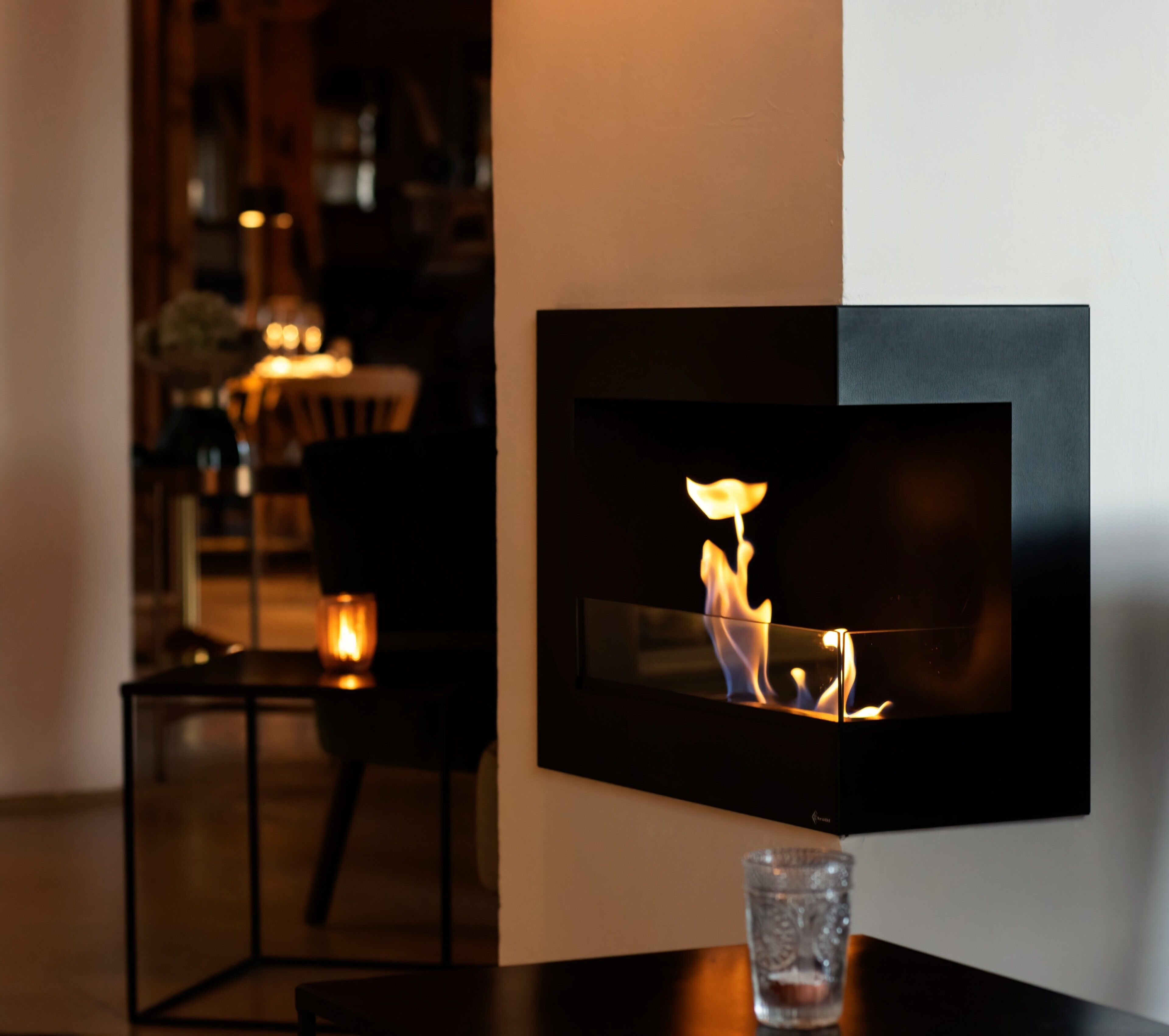 Un intérieur moderne avec une cheminée fixée au mur, une table en verre et une bougie posée sur une petite table d’appoint.